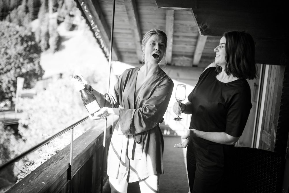 Black and white photo of bride in robe laughing with bridesmaid on sunlit wooden balcony.