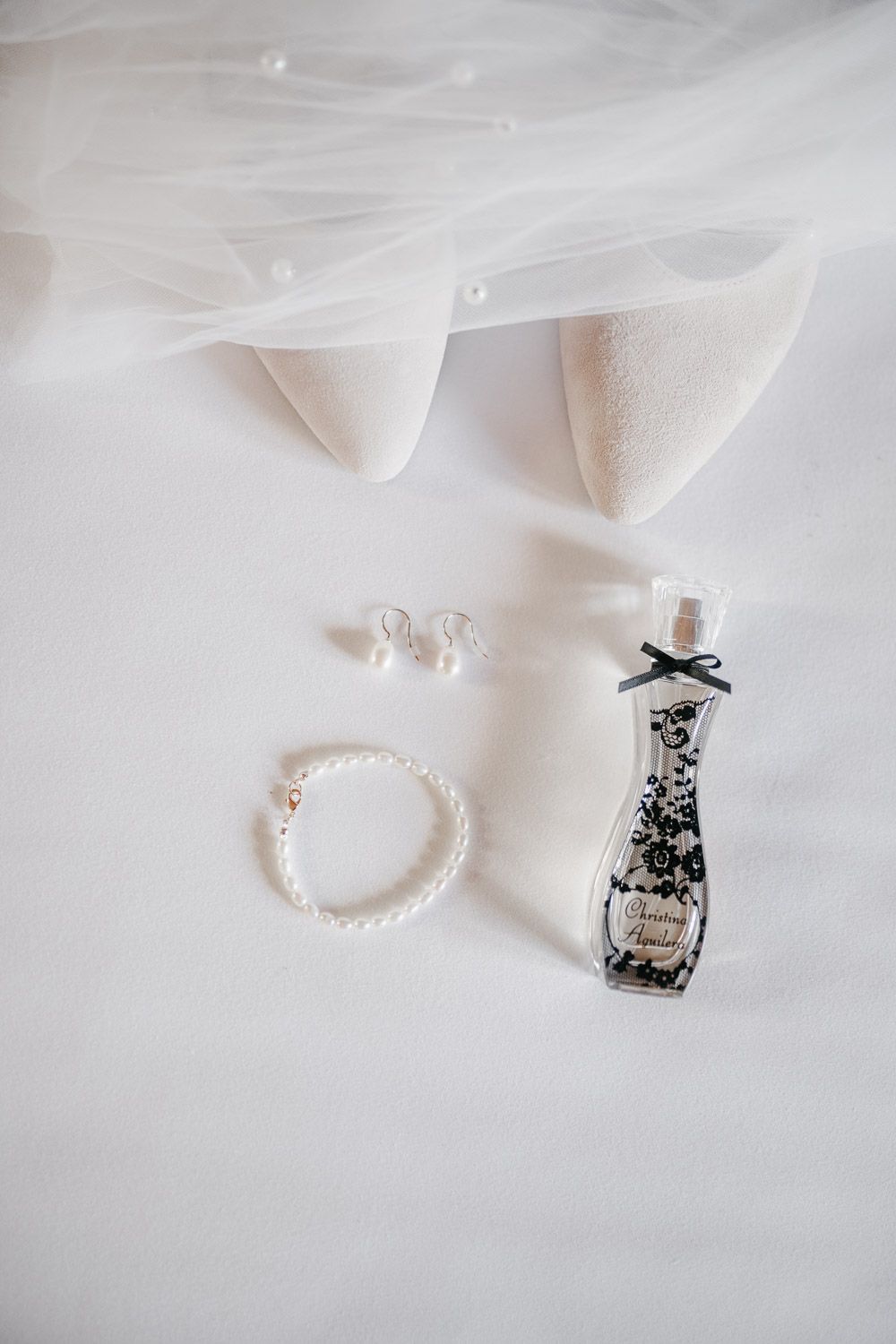 Bridal accessories flat lay featuring pearl bracelet, stud earrings, perfume bottle, and white heeled shoes.