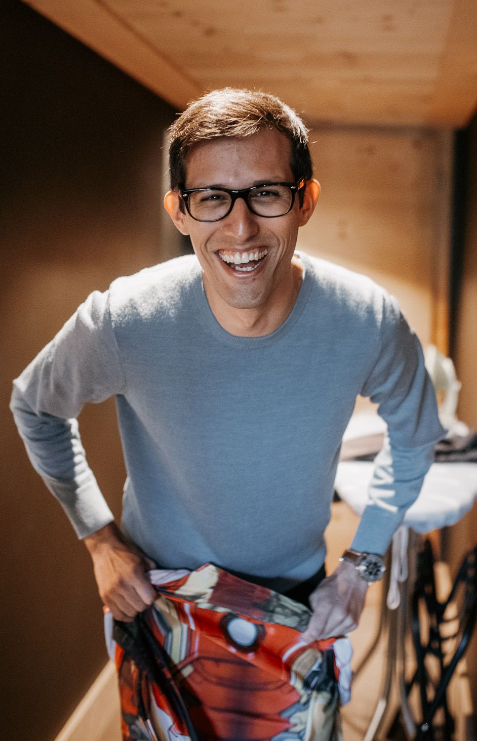 Smiling man in glasses and light blue sweater holding colorful shorts in a warmly lit wooden room.