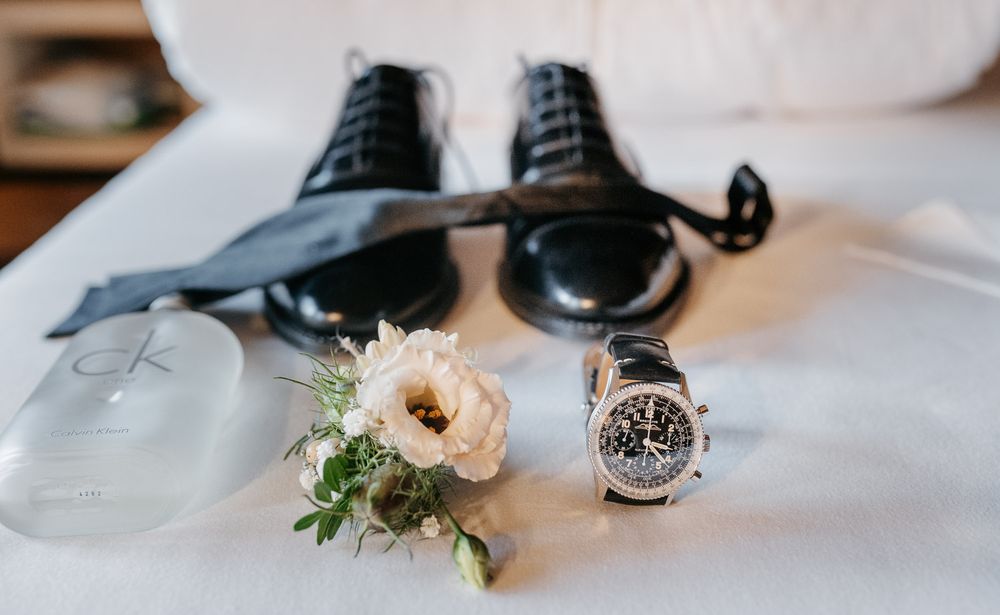 Wedding flat lay with black dress shoes, bow tie, boutonniere, watch, and Calvin Klein cologne on white surface.