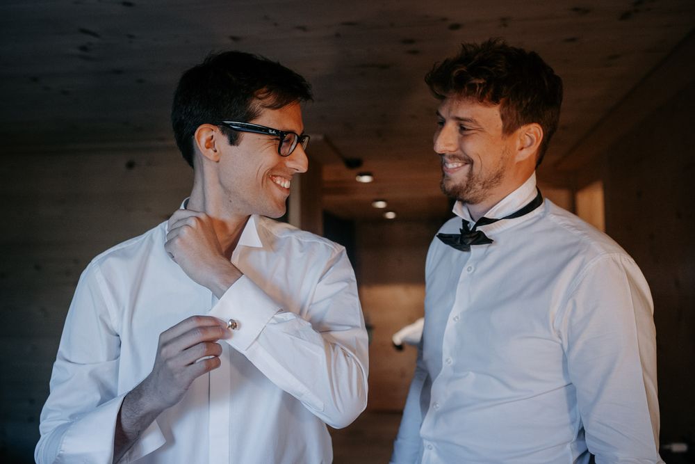 Two men in white dress shirts smiling while helping each other with cufflinks before a wedding.