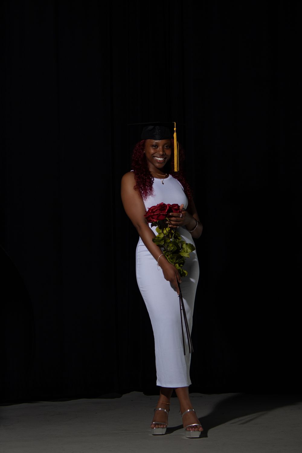 Graduate in elegant white dress holds red roses against dark backdrop.