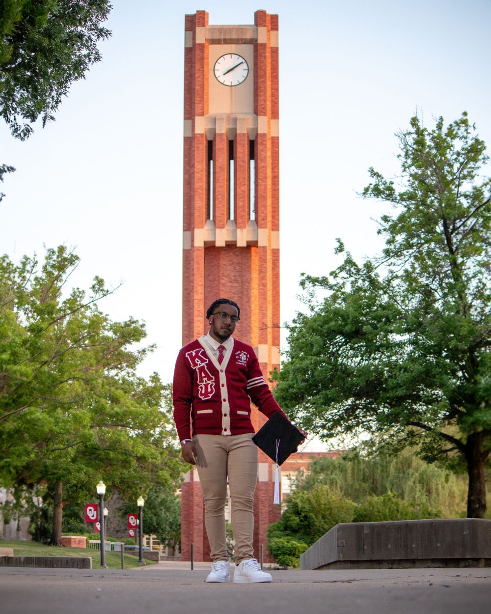 A figure poses in front of a brick clock tower wearing a burgundy cardigan and khaki pants.