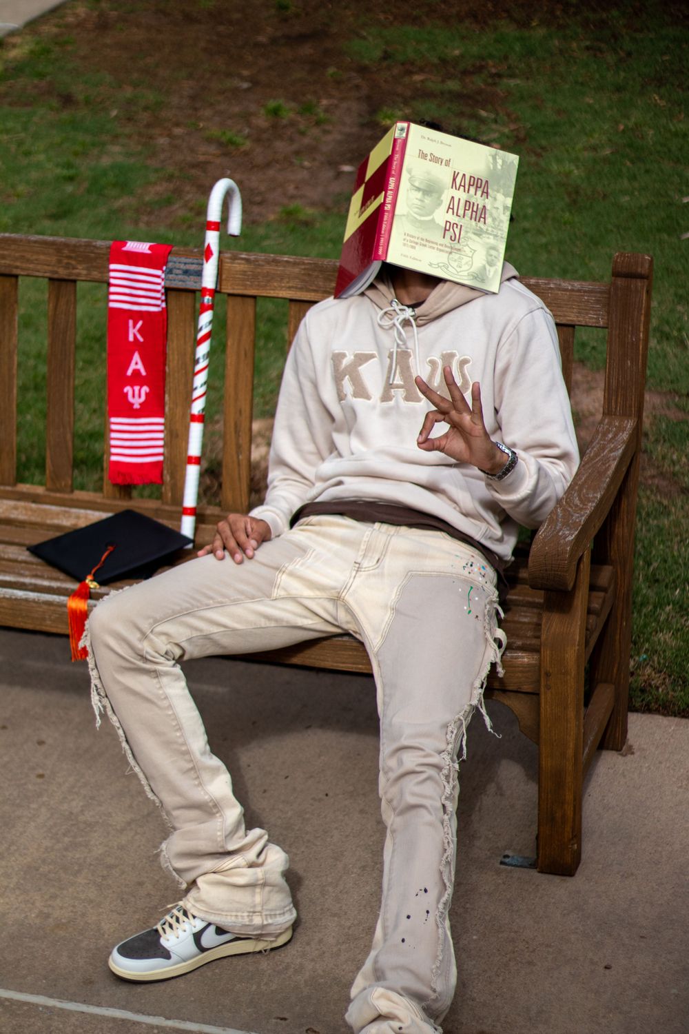 A person in light colored clothing sits on a bench with fraternity accessories.