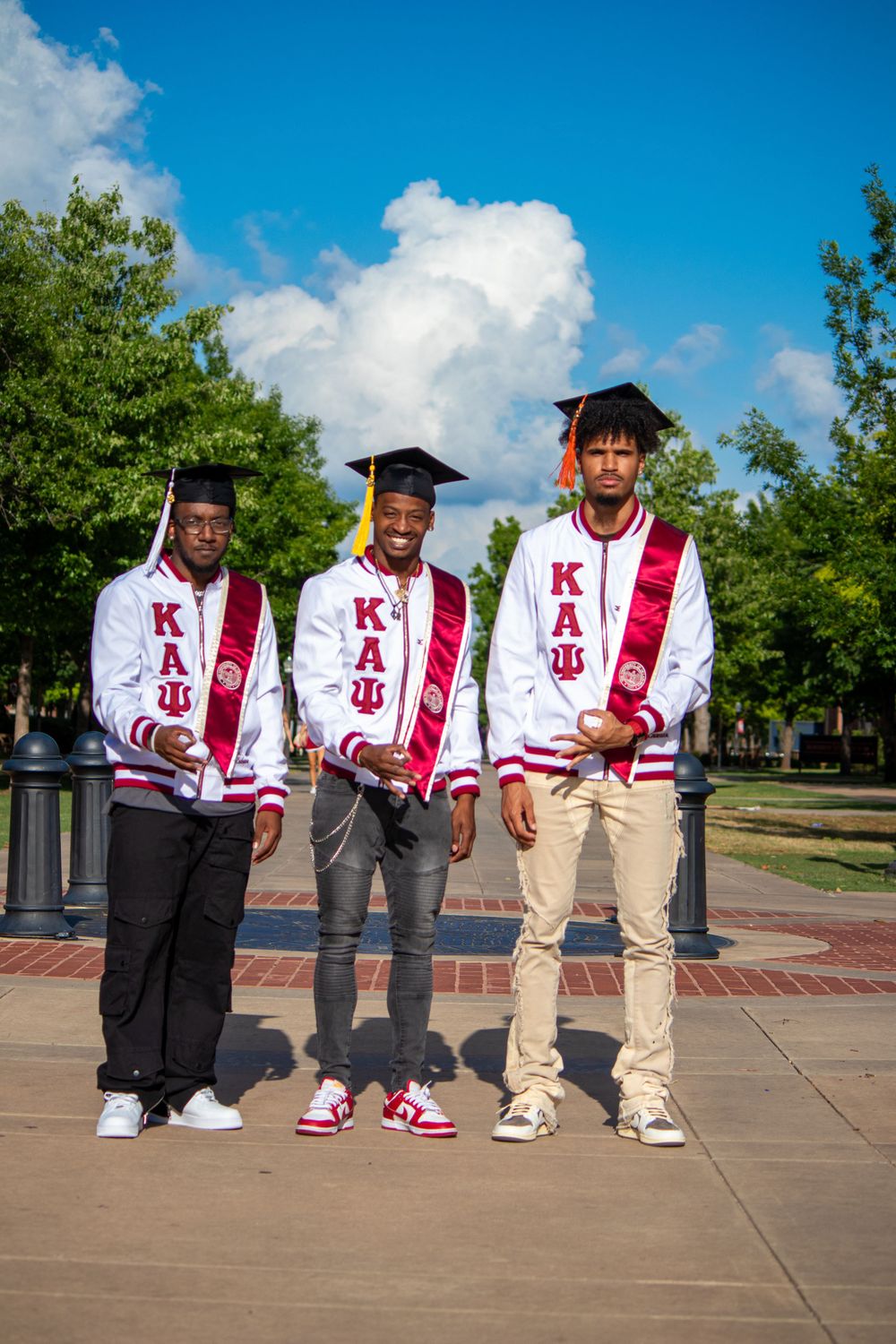 Three graduates in white and red stoles pose together on a sunny day.