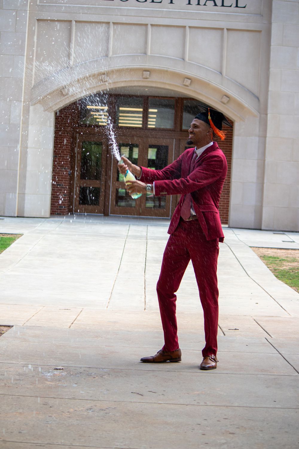 A graduate in a burgundy suit celebrates with confetti in front of a university building.