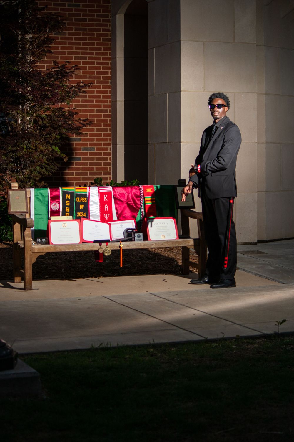 A person in formal wear stands next to a bench displaying colorful academic regalia.