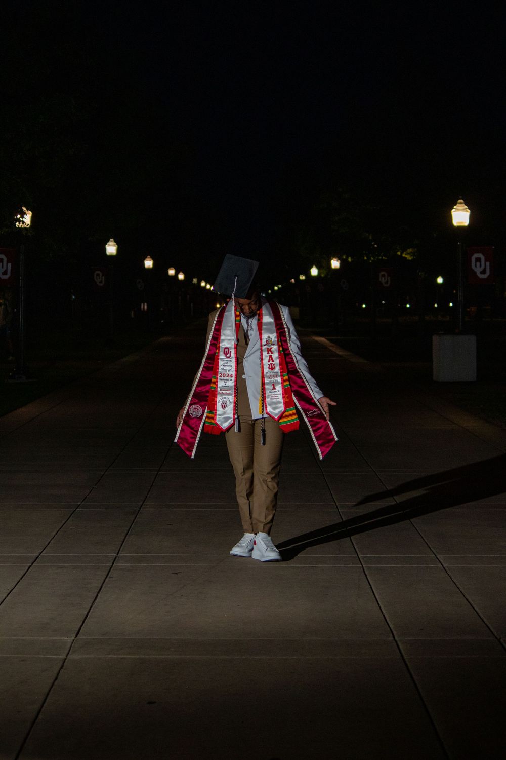 A silhouette with graduation stoles stands on a pathway lit by street lamps at night.