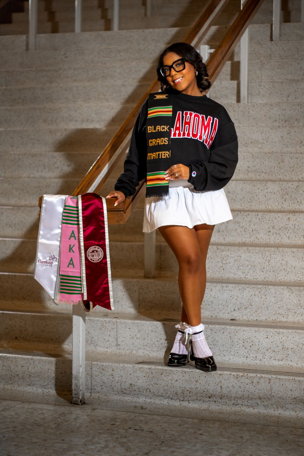 Student in AEOSU sweatshirt and white skirt poses on stairs with graduation stoles.