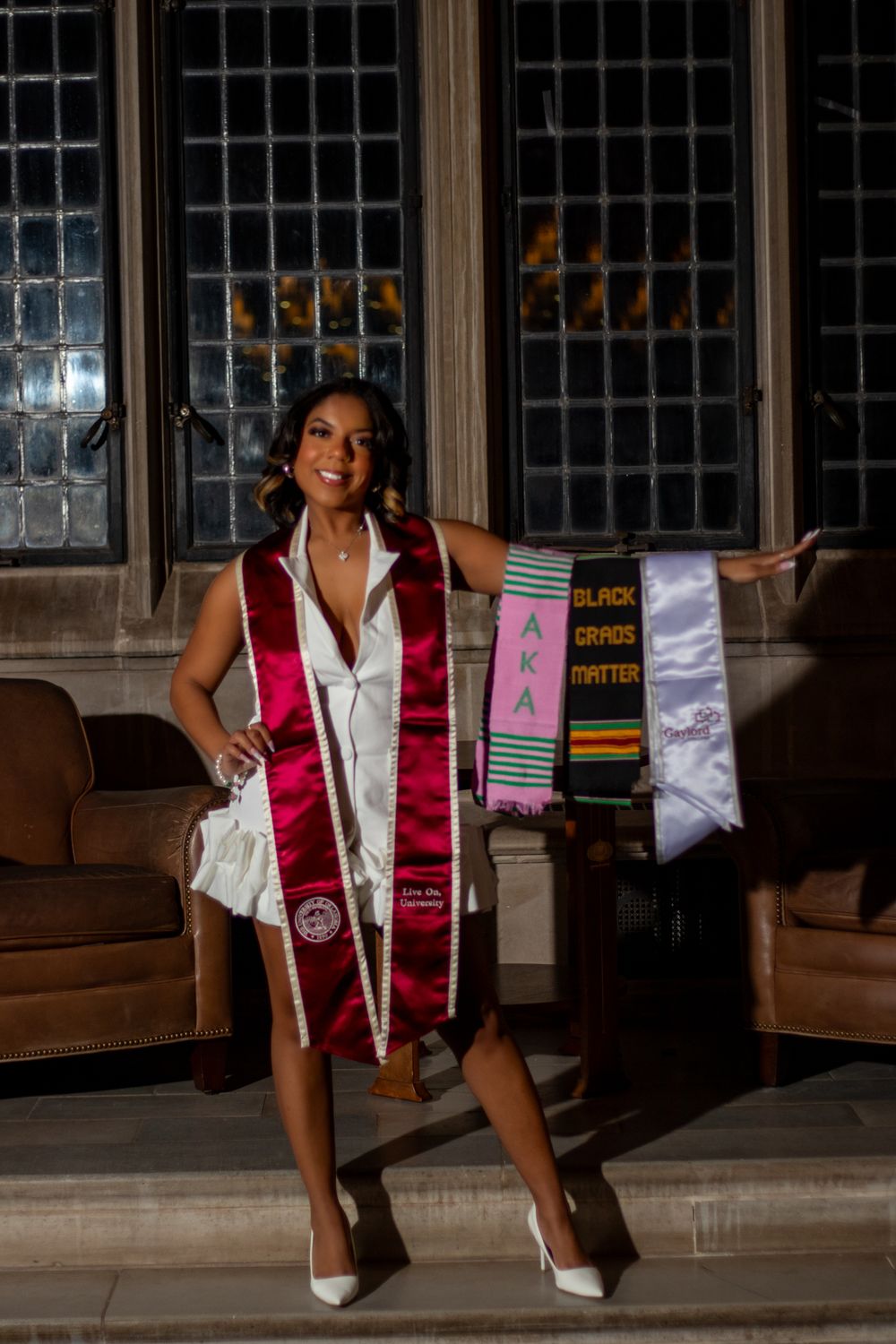 Graduate poses in white dress and maroon graduation stole near decorative banners.