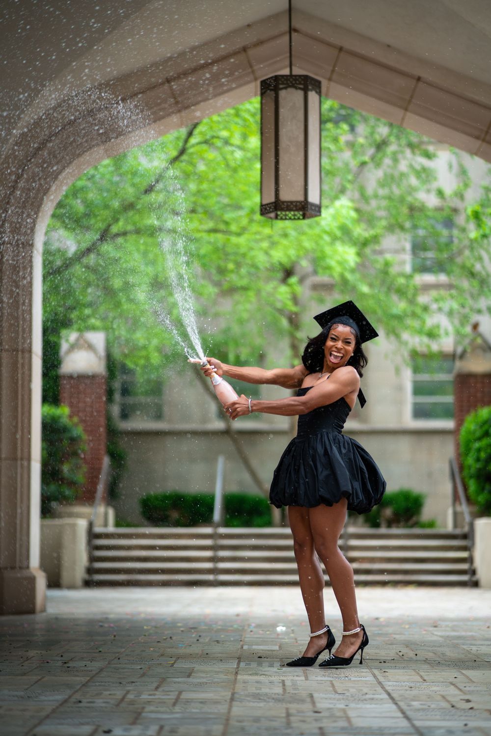 Graduate in black dress and cap celebrates with champagne spray under outdoor archway on graduation day.