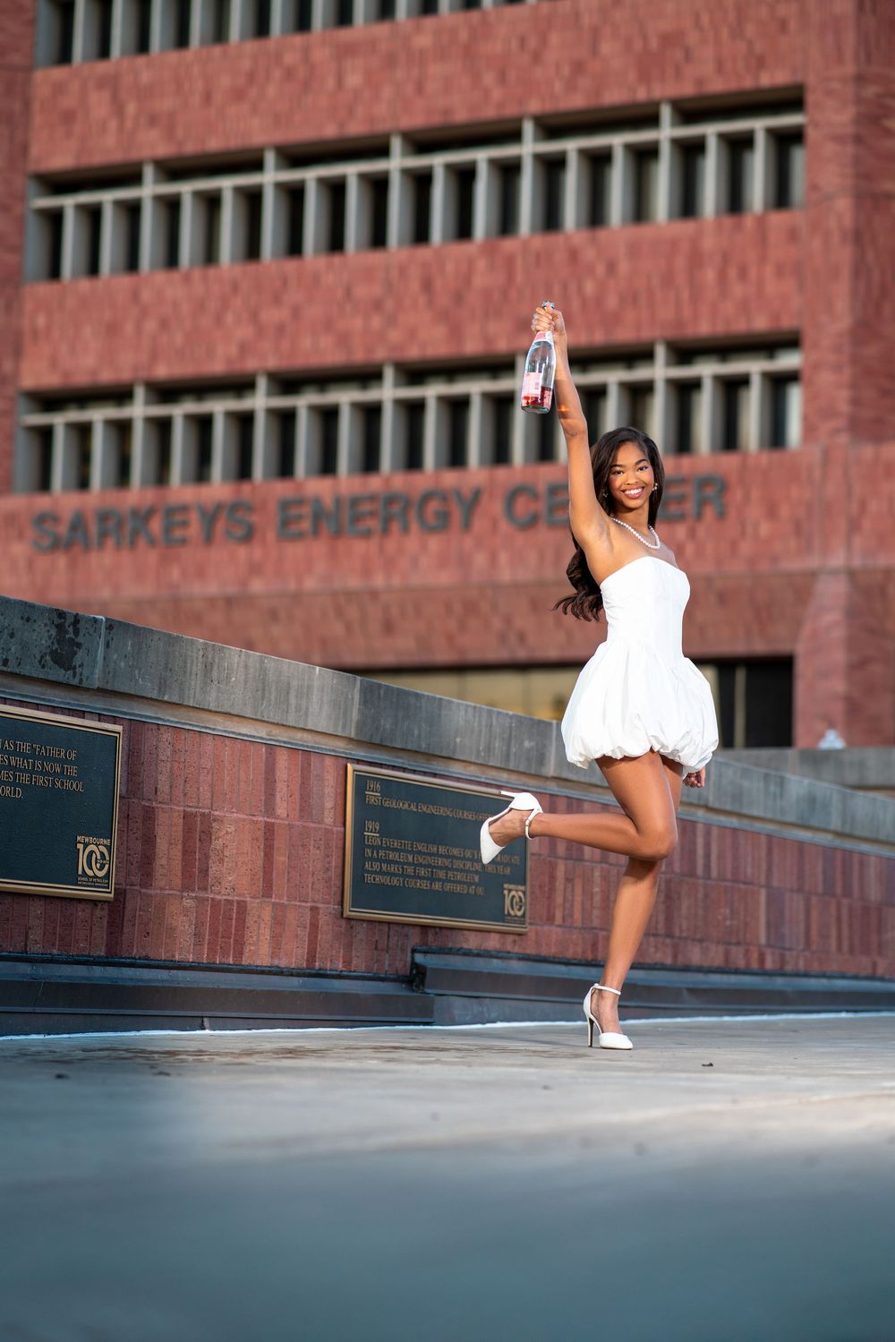 A graduate celebrates outside Sarkeys Energy Center in a white dress and high heels.