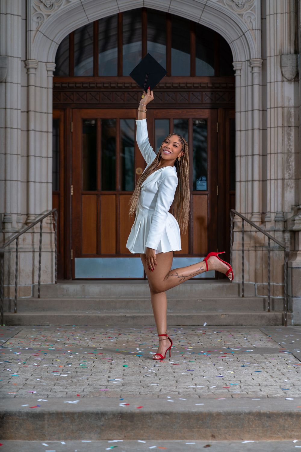 Graduate celebrates with graduation cap and red heels while posing playfully in front of ornate university doorway.