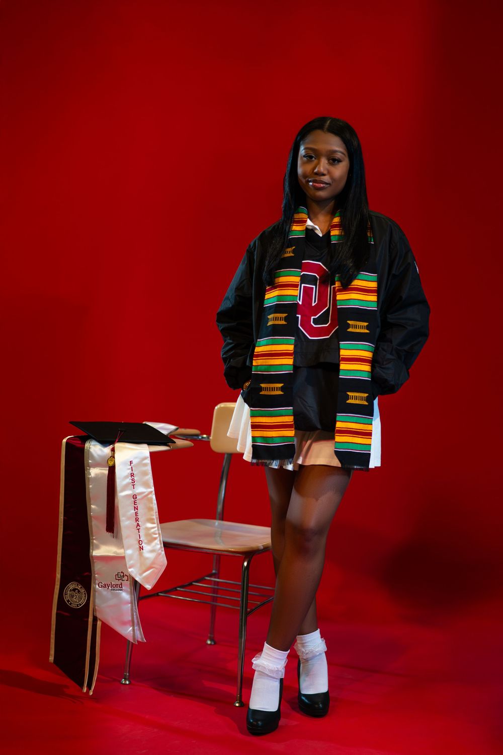 Graduate posing in black graduation attire with colorful kente stole against a red backdrop.