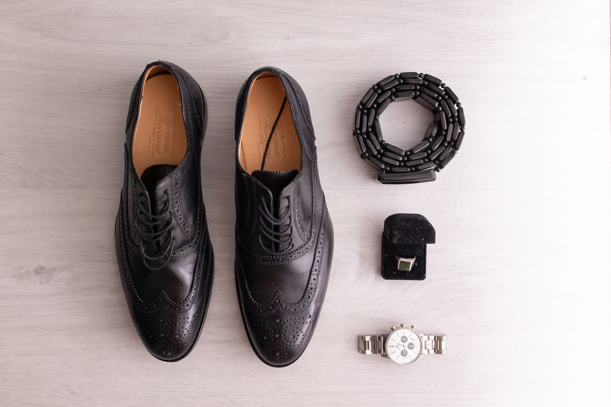 Elegant black wingtip dress shoes arranged with a belt, watch and ring on gray surface.