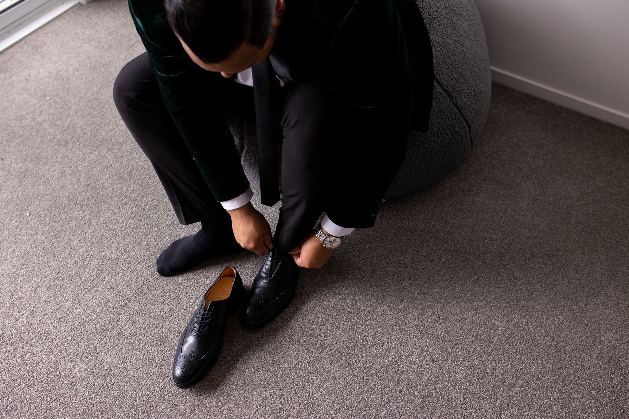 Person putting on formal black leather dress shoes while sitting on gray carpet.