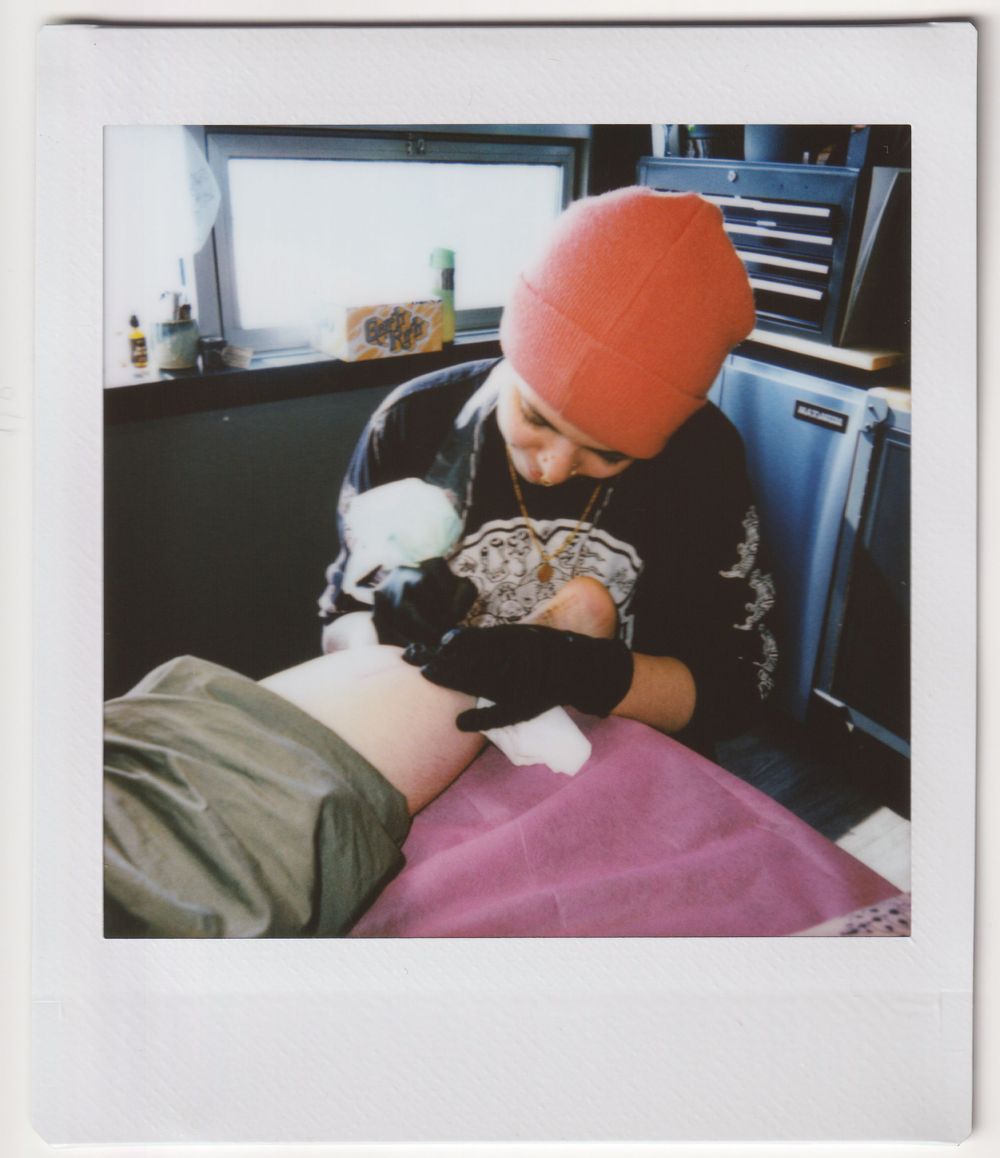 Tattoo artist wearing an orange beanie works on a client in a professional studio setting.