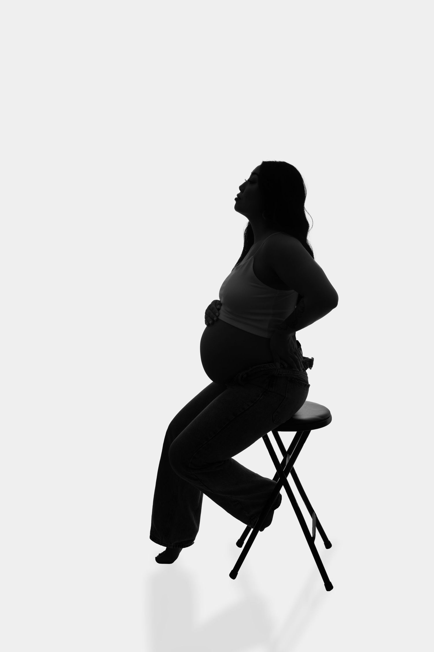 Silhouette of pregnant woman sitting on stool against white backdrop.