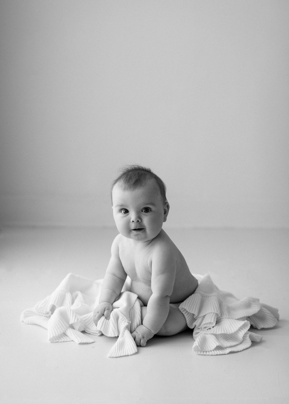 A baby in black and white sits on white fabric while looking towards the camera with a sweet expression.