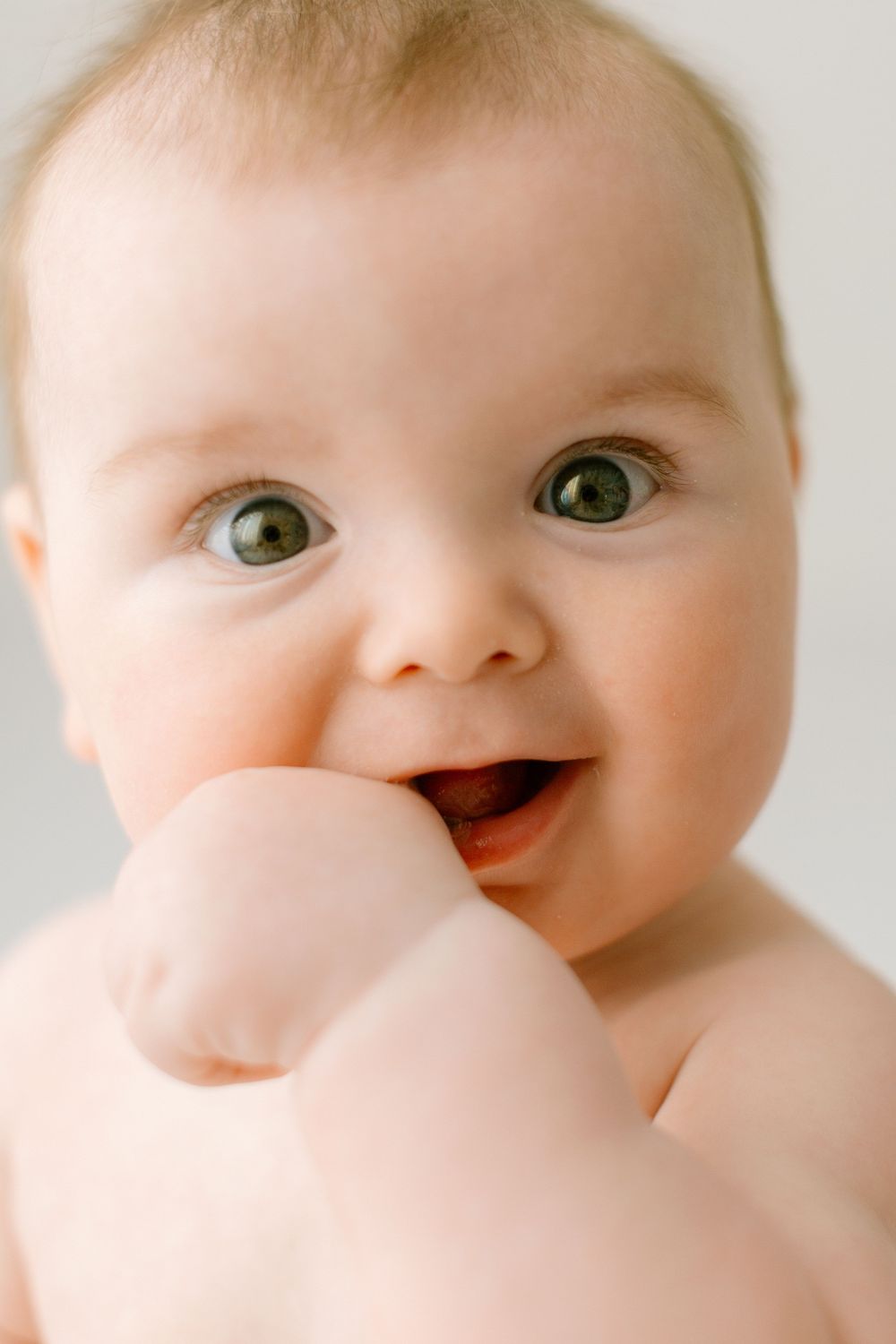 A happy baby with bright green eyes smiles while putting their hand near their mouth.