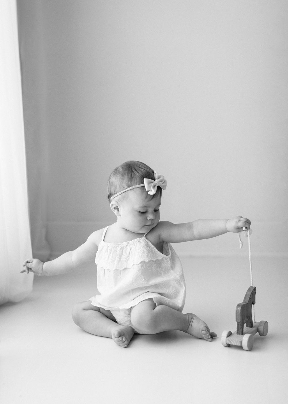 A sweet baby in white romper with bow headband plays with a wooden pull toy in black and white photograph.