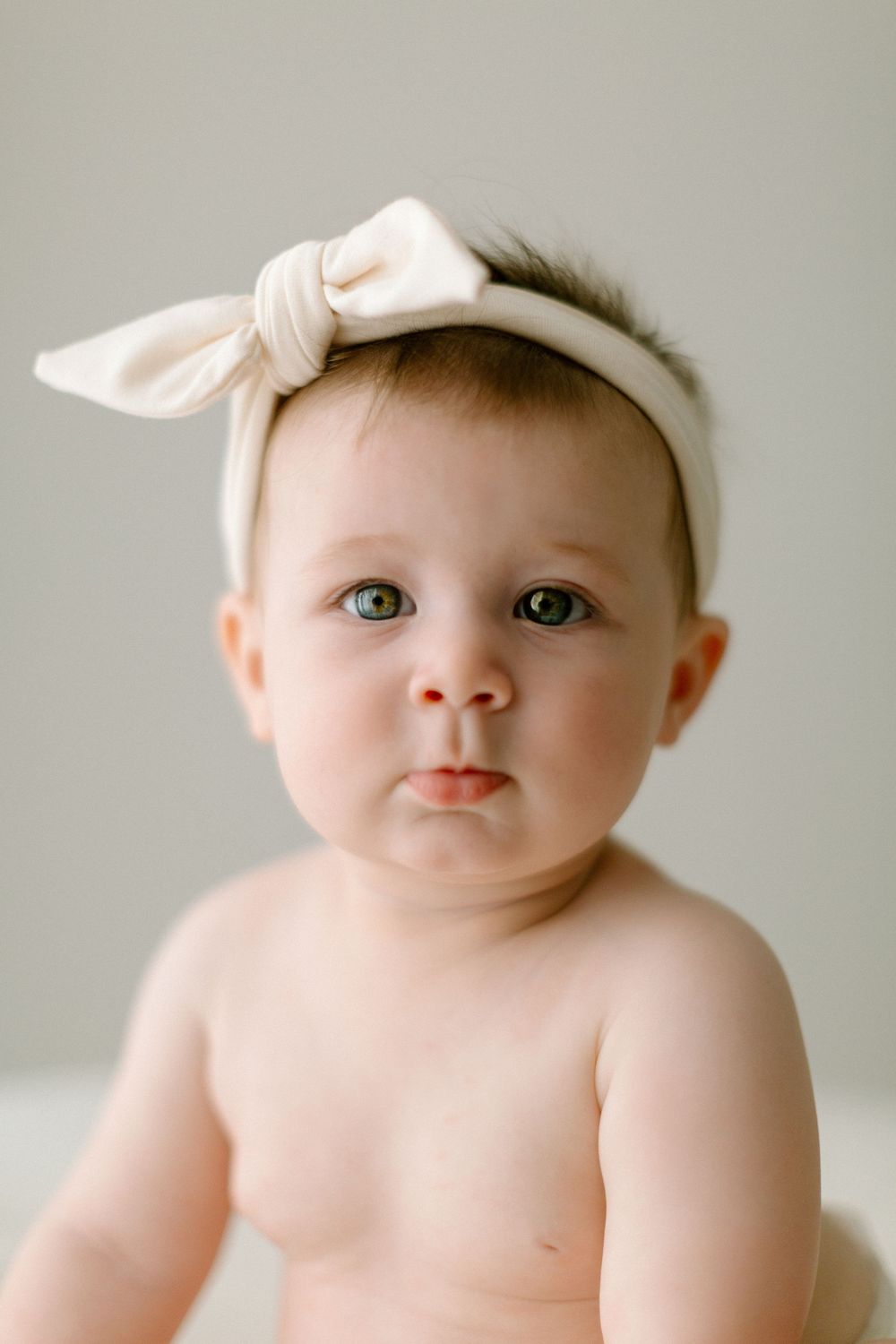 Close up portrait of a baby wearing a white bow headband against a neutral gray background.