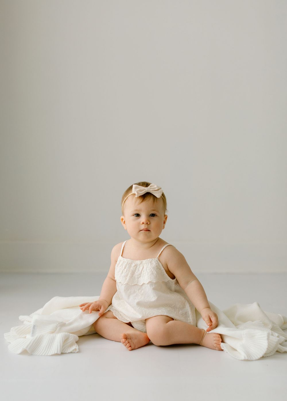 Baby in a white sleeveless romper sits on light fabric against neutral background.
