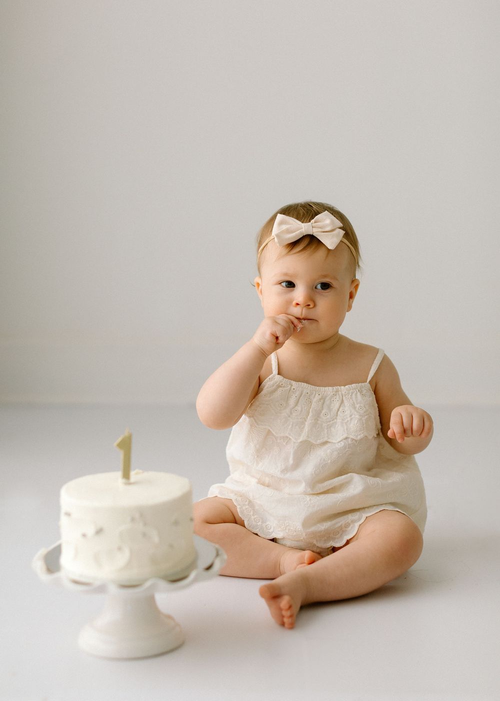 Baby in white outfit sits beside small white birthday cake with number one candle.