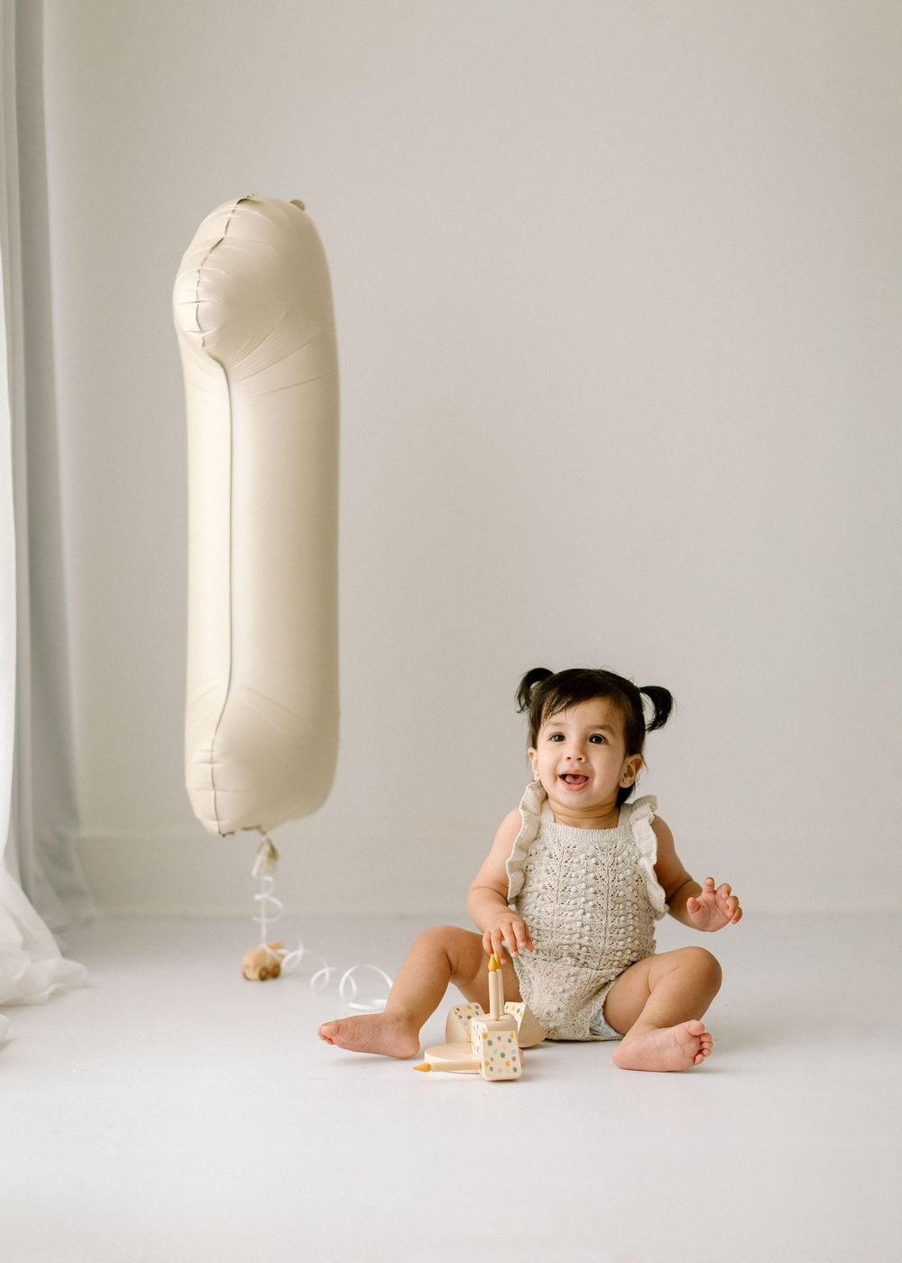 Toddler in beige knit romper sits on white floor with wooden toy and large white balloon floating nearby.