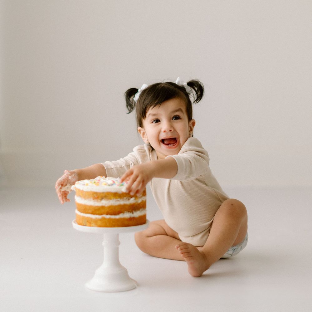 A feminine figure in beige tones interacts with a layered white cake on a cake stand against a neutral background.