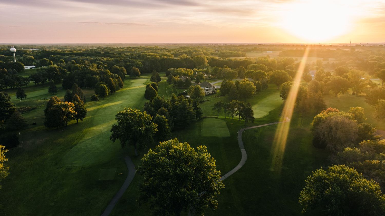 Aerial view of a golf course at sunset with winding paths and lush green trees casting long shadows.