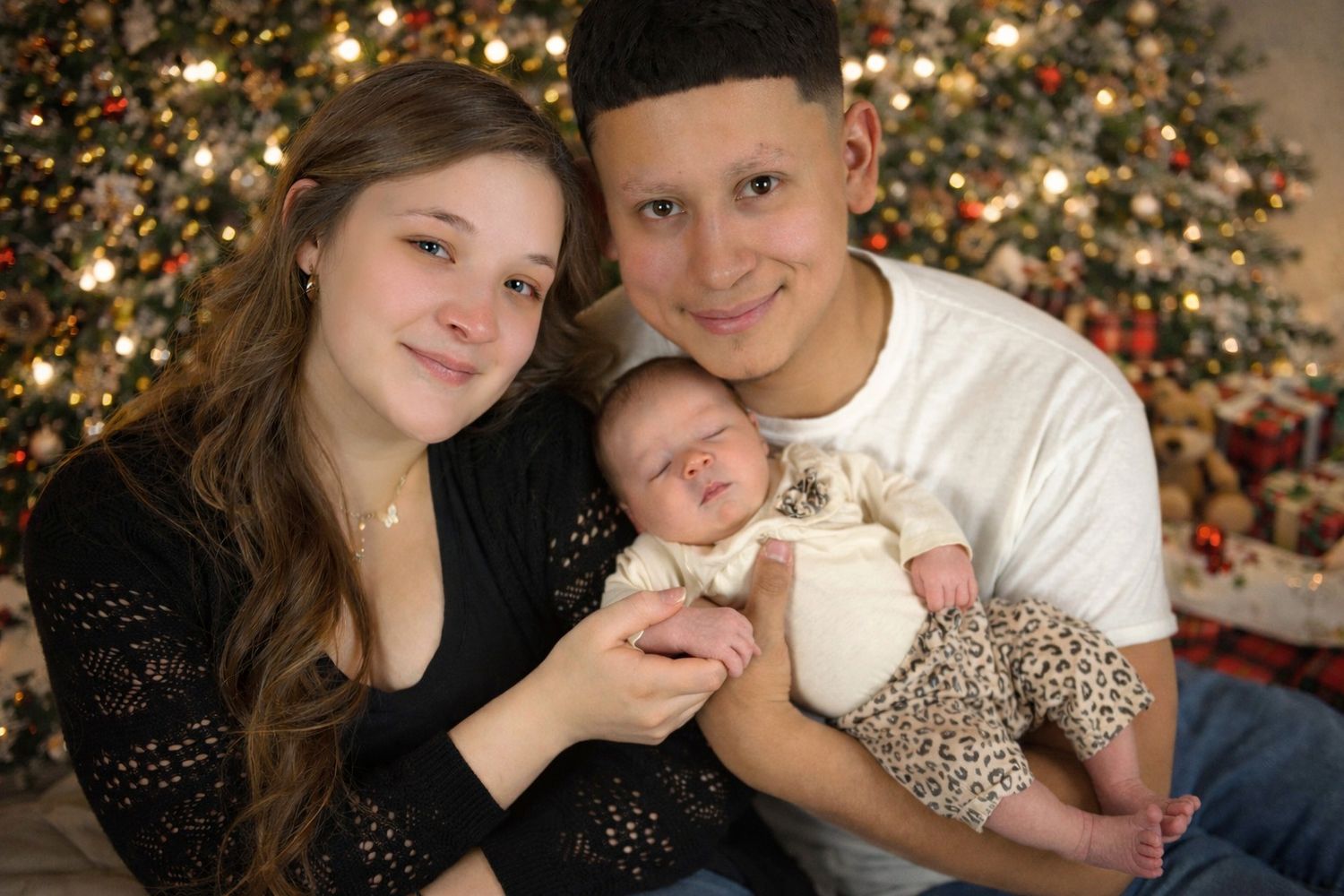 Family poses together near Christmas tree with twinkling lights creating a warm holiday atmosphere.