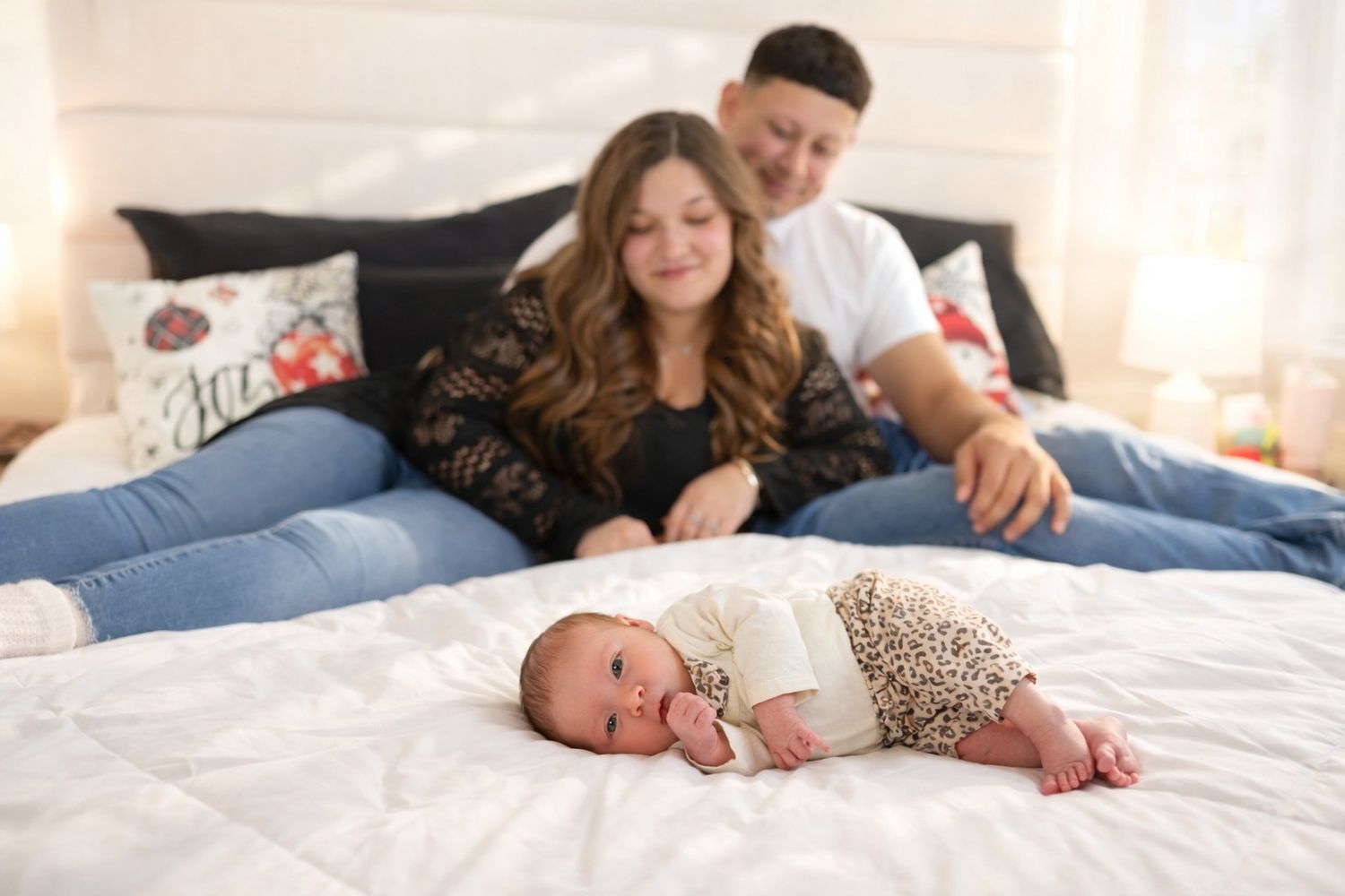 A baby in leopard print pants lies on a white bed while two people relax behind on pillows.