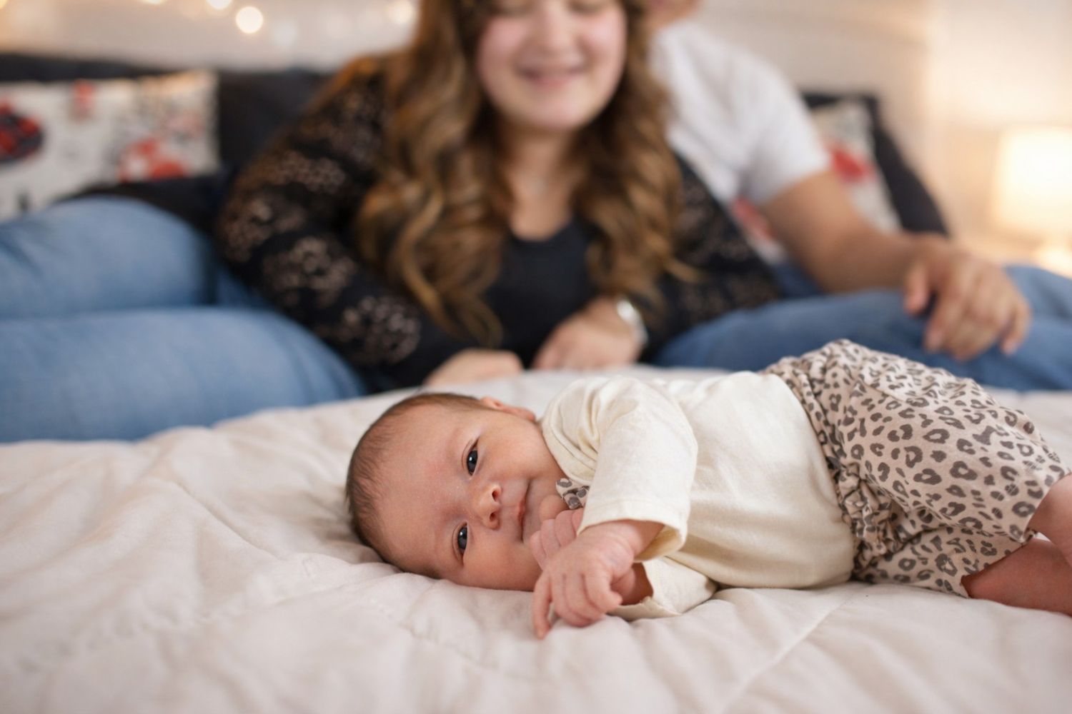 Baby in white outfit lies on soft bedding while looking at camera with a gentle smile.
