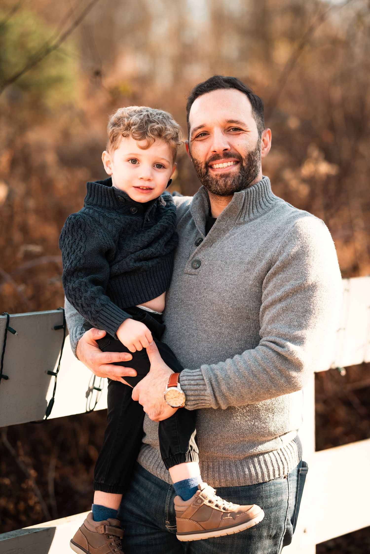 A parent and child share tender moments together while sitting on a white fence in an autumn outdoor setting.