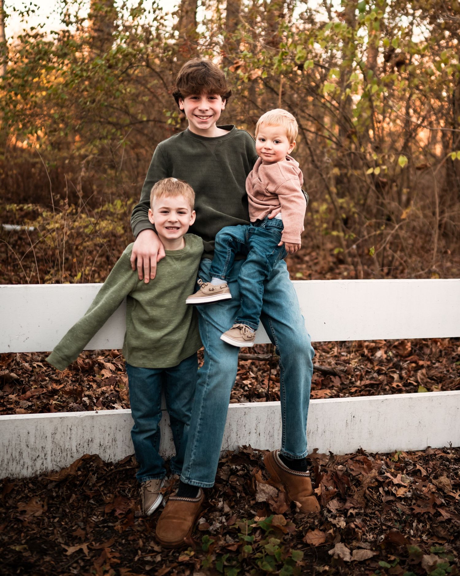 A family sits together on a white fence surrounded by fallen autumn leaves in a wooded setting.