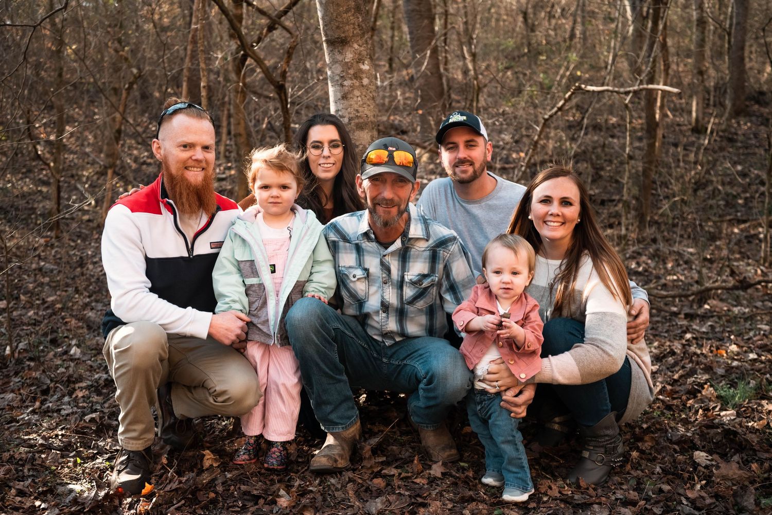 Extended family gathering poses together outdoors in autumn woodland setting.