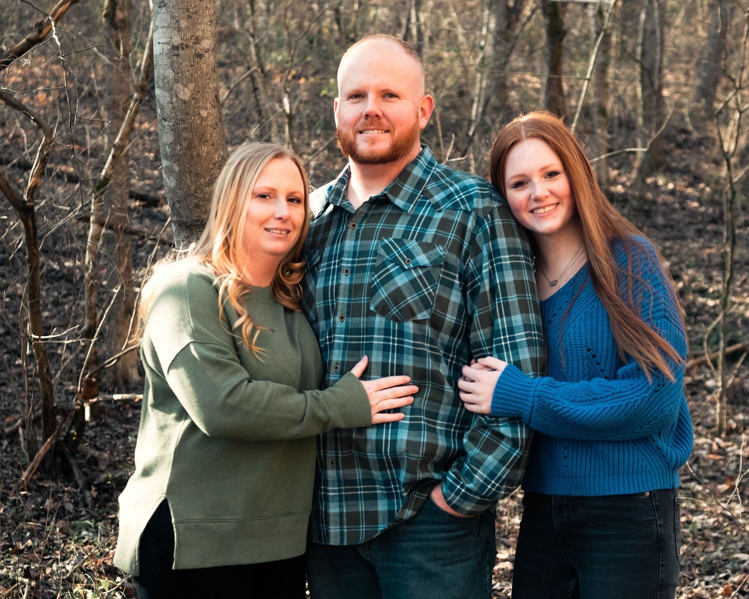 Three people pose together in casual wear against a winter forest backdrop.