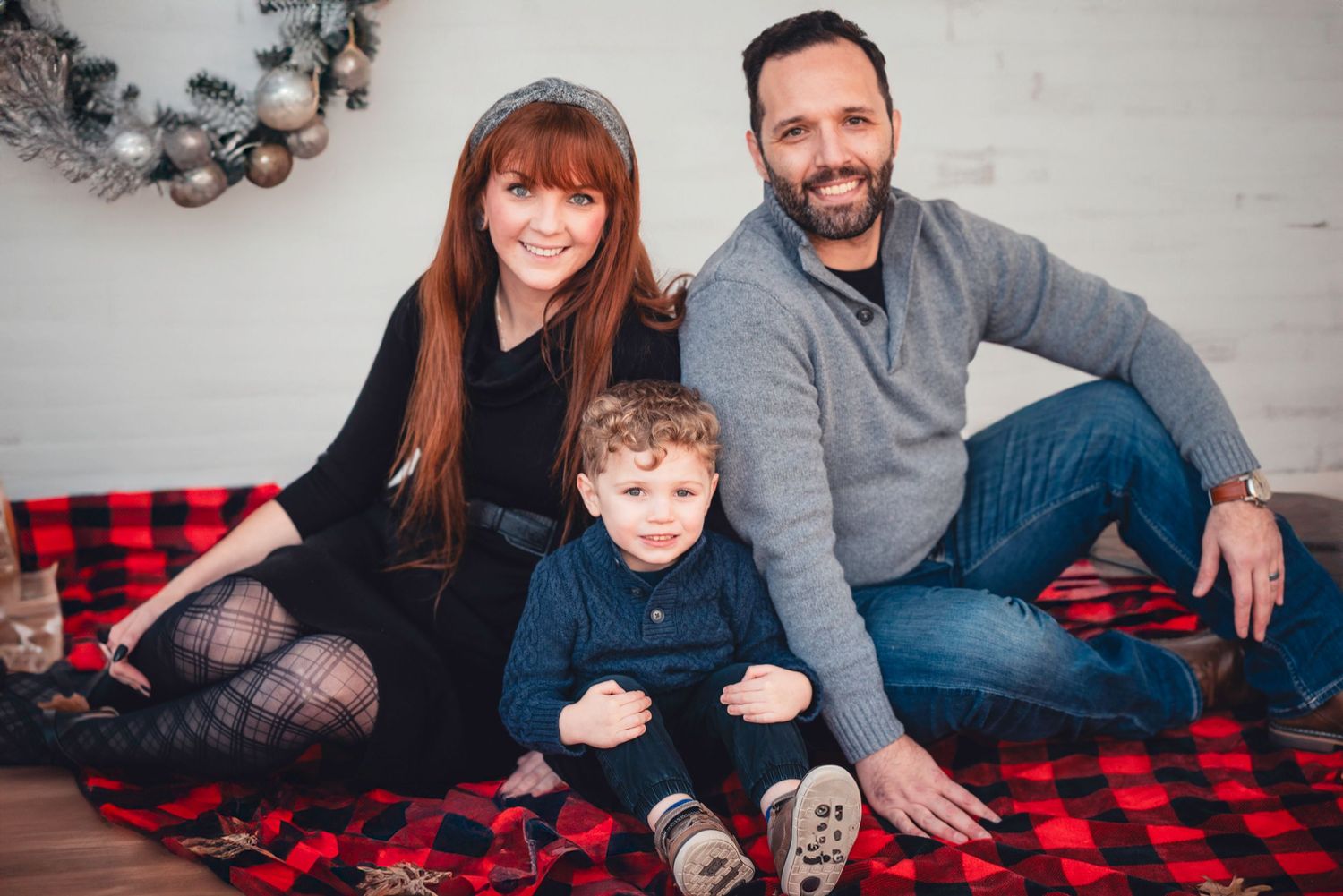 A family sits together on a red and black buffalo plaid blanket for a casual indoor portrait session.