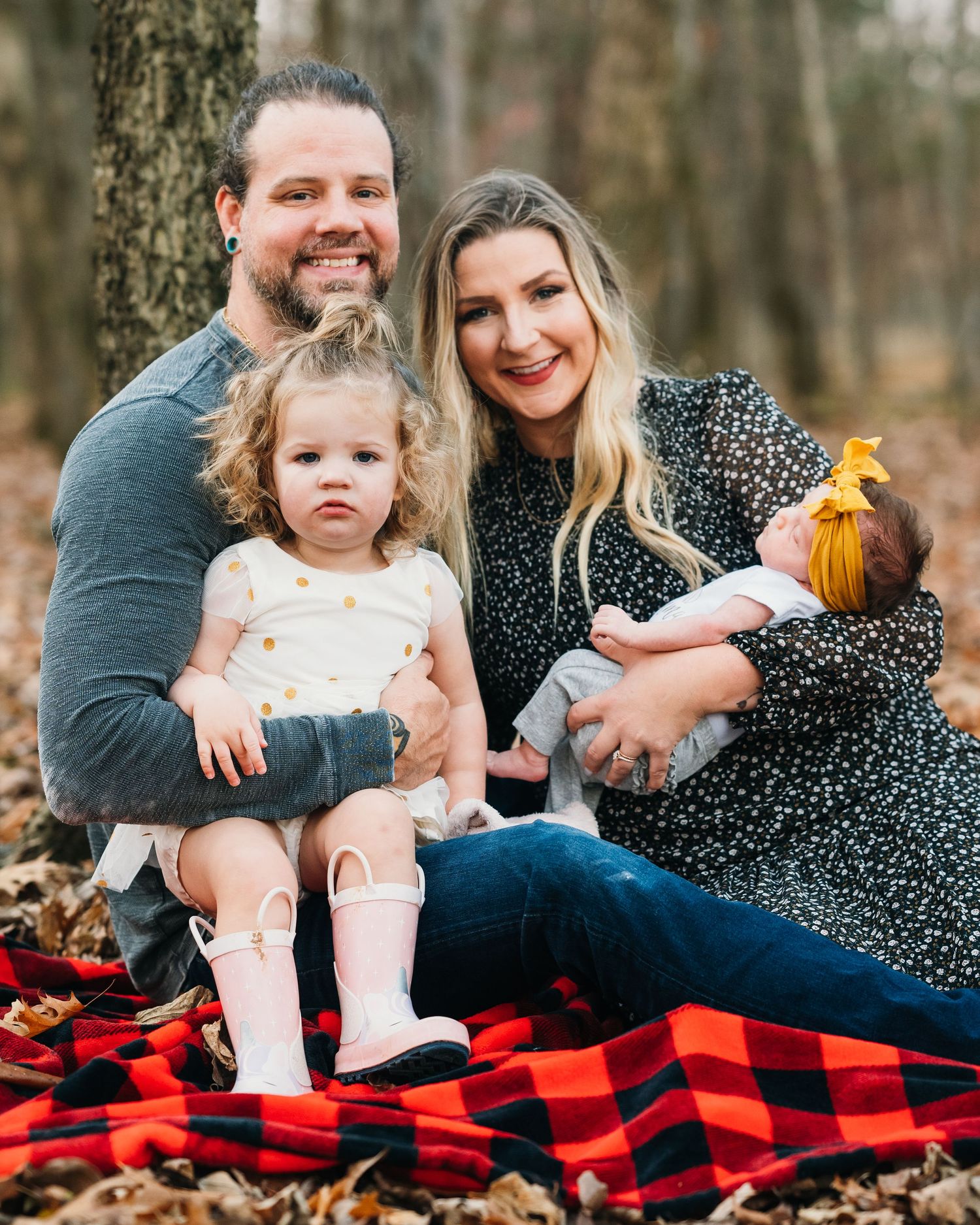 A family sits together on a red and black buffalo plaid blanket during an outdoor photo session in nature.