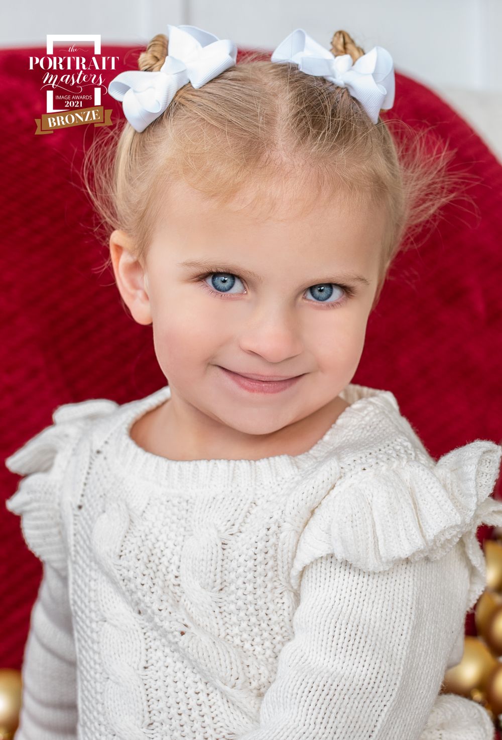 Portrait of young child in white knit sweater with white bows against red background.