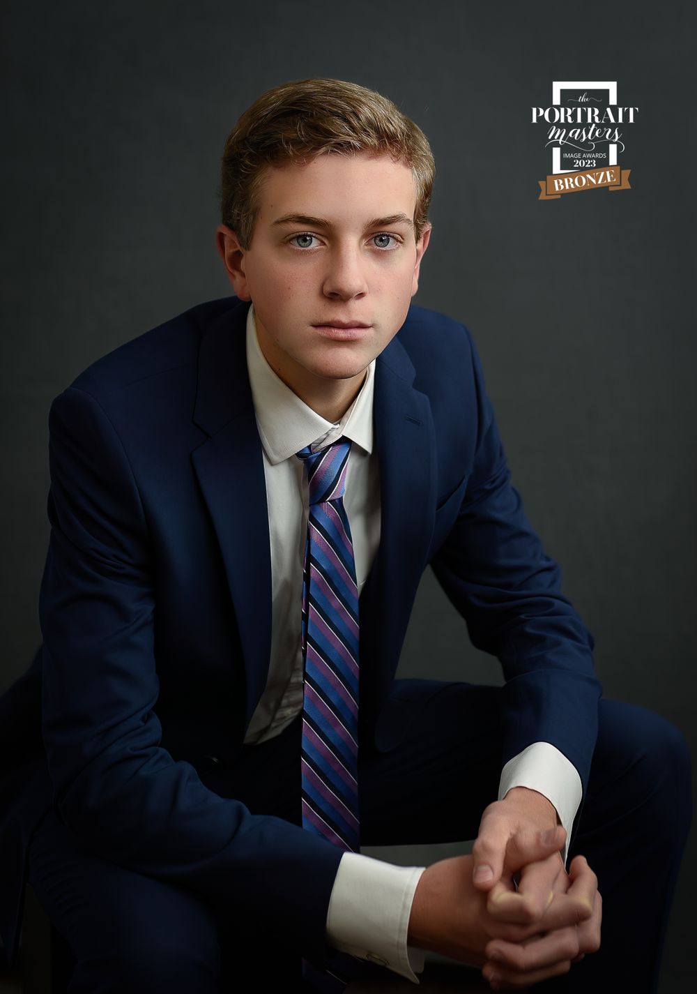 Formal portrait in navy suit with striped tie against dark background.