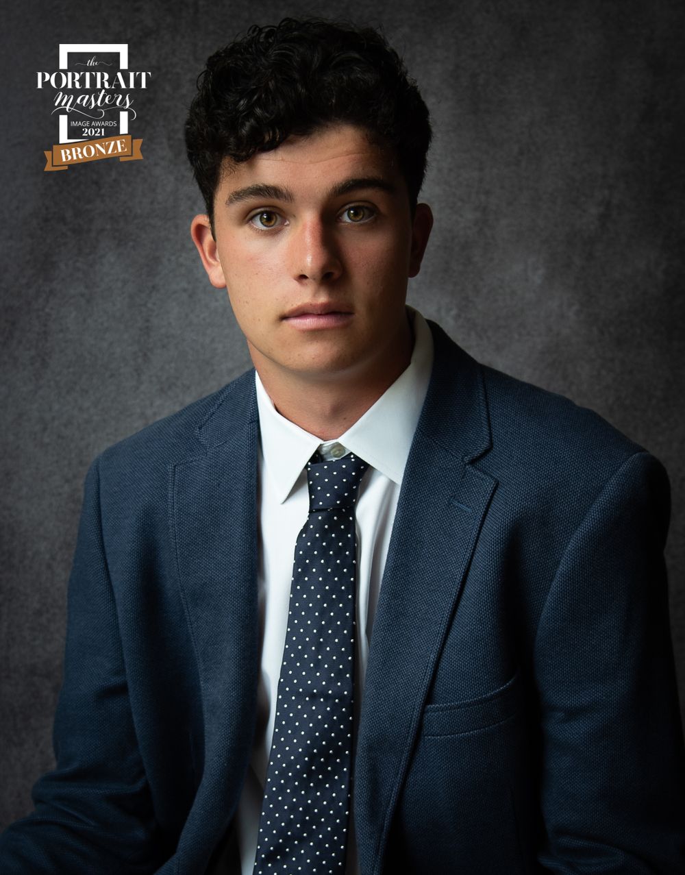 Professional headshot in blue suit with polka dot tie against textured background.