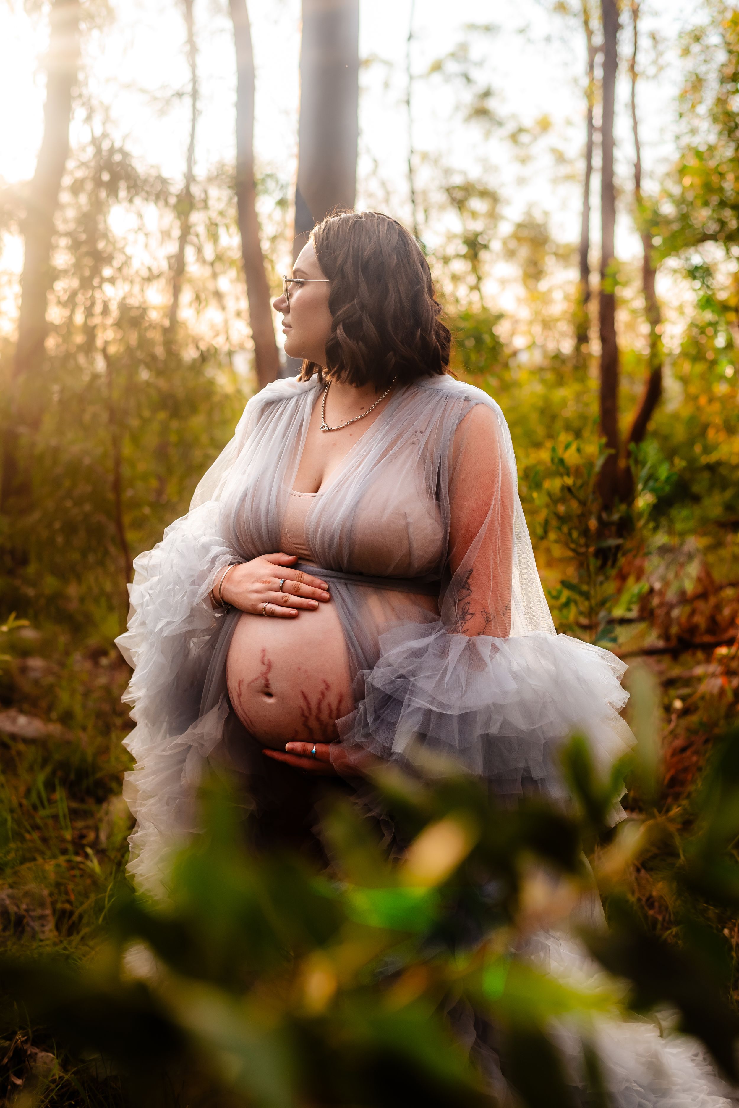 An ethereal maternity photo in a sunlit forest featuring a flowing tulle dress.