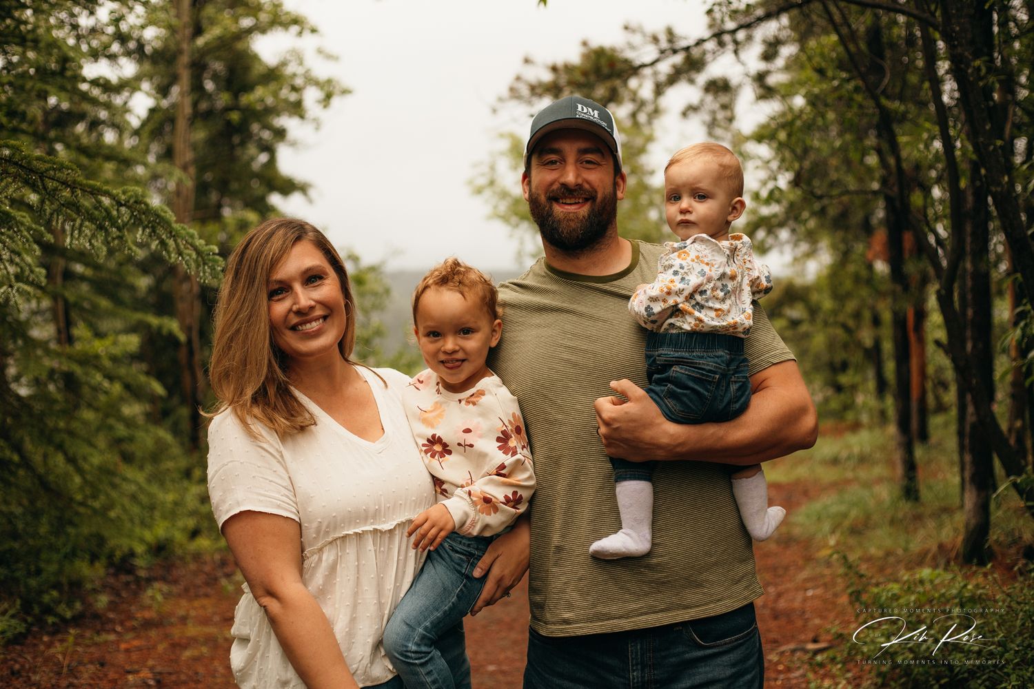 Smiling family of four with two young children posing on a forest trail.