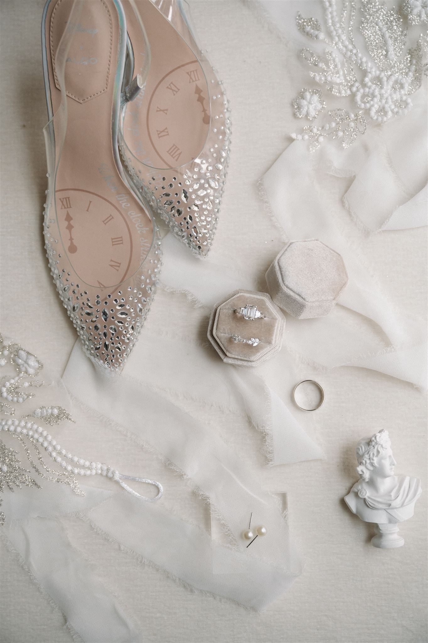 Wedding details including crystal-embellished shoes, rings, and pearls arranged on white fabric.