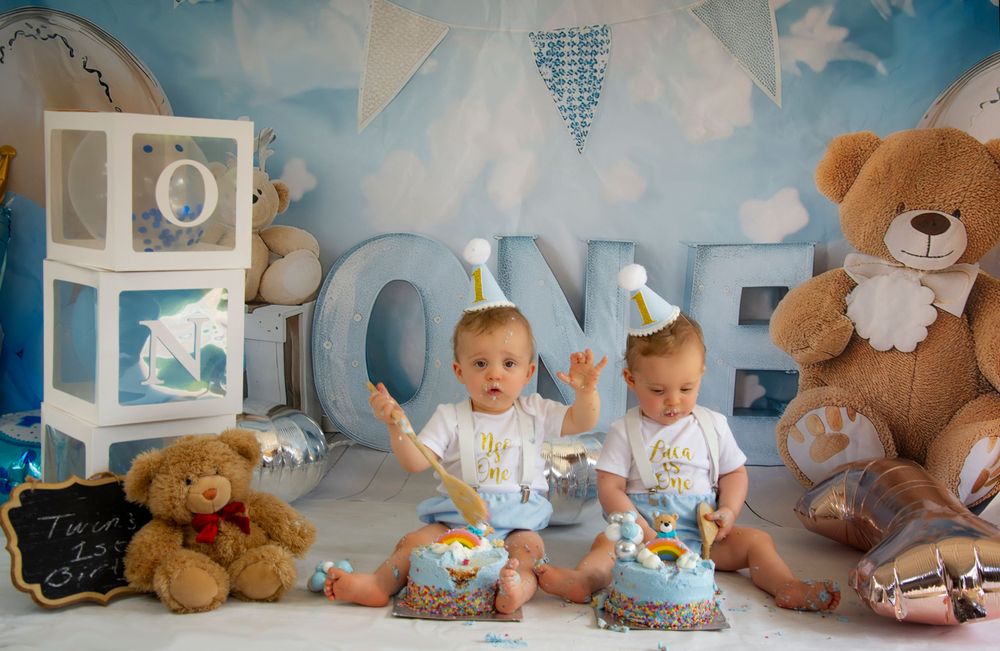 Twin babies in matching outfits sit with cake smash props and teddy bears against a blue ONE backdrop.