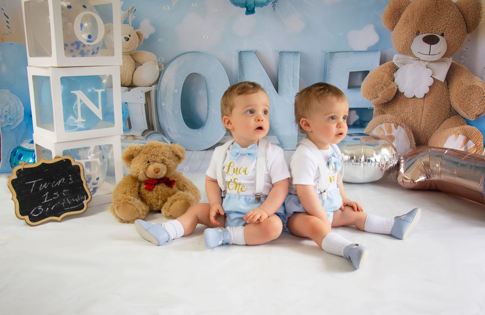 Twin babies sitting together against blue and white ONE letter decorations with teddy bears.