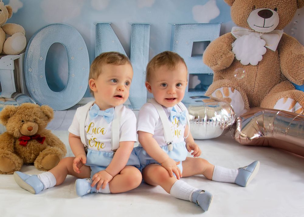 Twin babies in matching outfits sitting together for first birthday photoshoot with blue decor.
