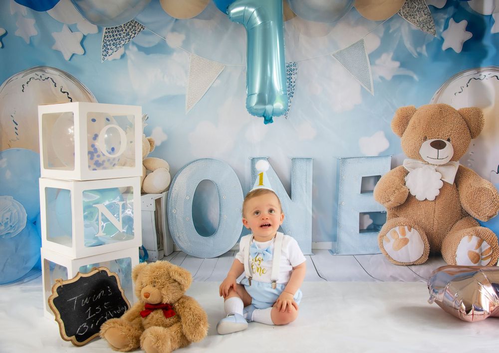 Baby sitting beside decorative boxes and teddy bears with blue ONE letters in background.